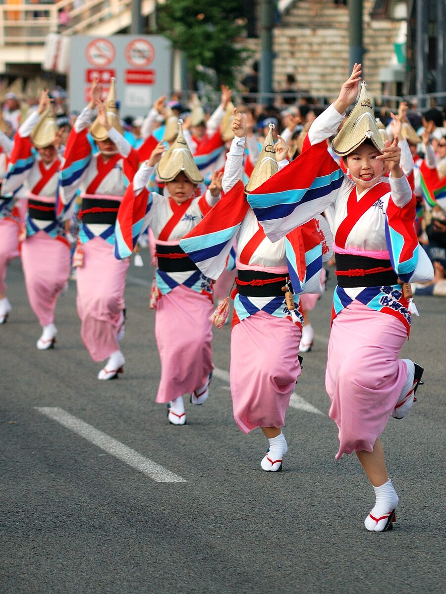 Fila di danzatrici dell’Awa Odori in kimono rosa, giacche rosse e amigasa, con le braccia alte durante l’avanzamento.
