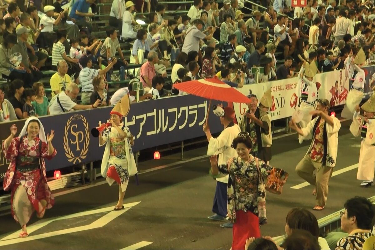 Danzatori del Muso-ren avanzano in una strada gremita di Tokushima; una danzatrice regge un parasole rosso davanti alle tribune.