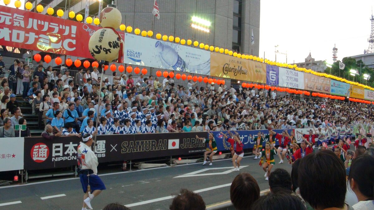 Veduta ampia dell’Awa Odori di Tokushima: il ren avanza lungo la strada davanti alle tribune, sotto file di lanterne.