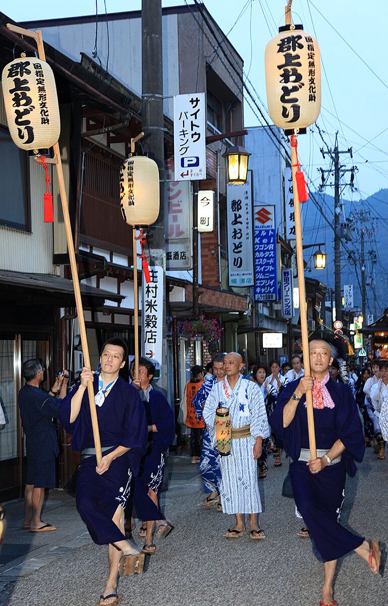 Due uomini in abito scuro avanzano su geta portando alte lanterne con il nome Gujō Odori, seguiti da partecipanti in yukata.