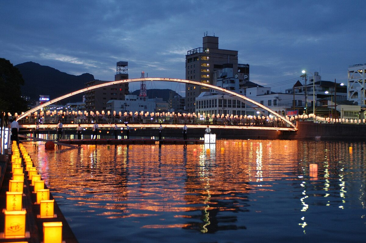 Lanterne illuminate galleggiano sul fiume Sasebo al crepuscolo, davanti a un ponte ad arco acceso.