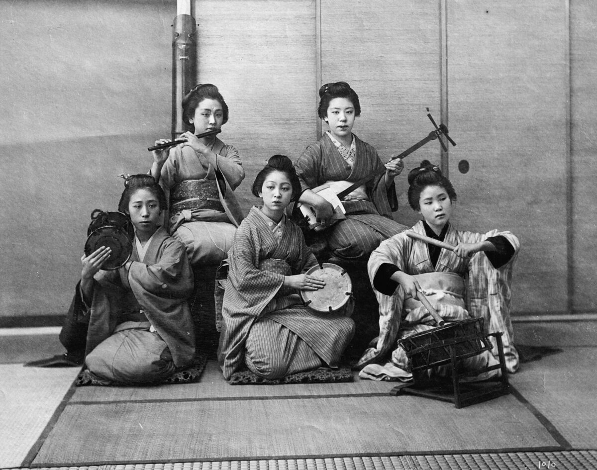 Fotografia storica in bianco e nero di un gruppo di musiciste giapponesi con shamisen, flauto e tamburi tradizionali.