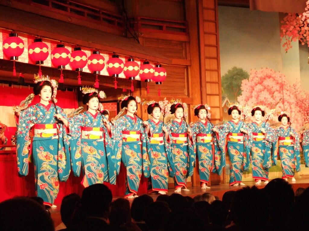 Gruppo di maiko in kimono turchese danza in fila sul palco del Miyako Odori, sotto lanterne rosse e una scena di ciliegi.