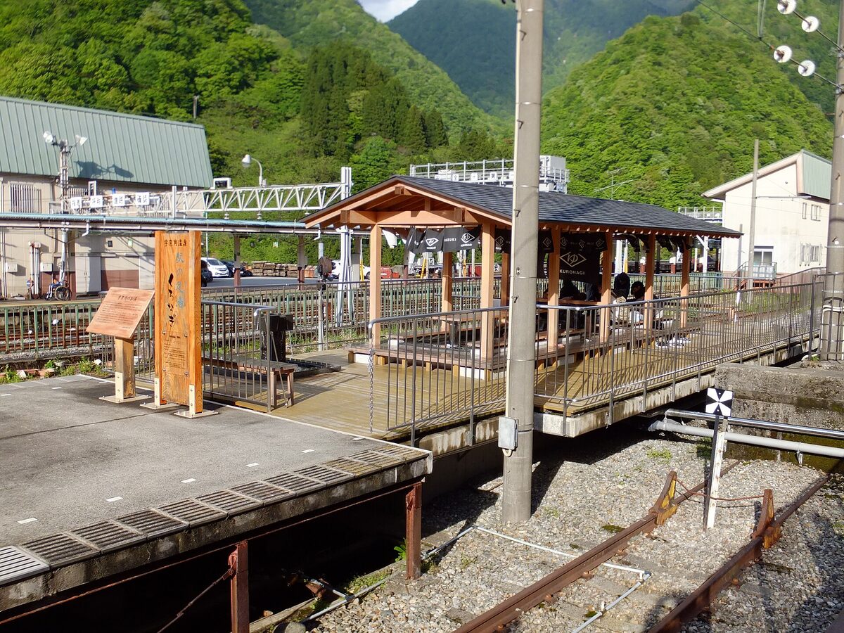 Padiglione in legno con vasca ashiyu accanto ai binari della stazione di Unazuki Onsen, fra montagne verdi.