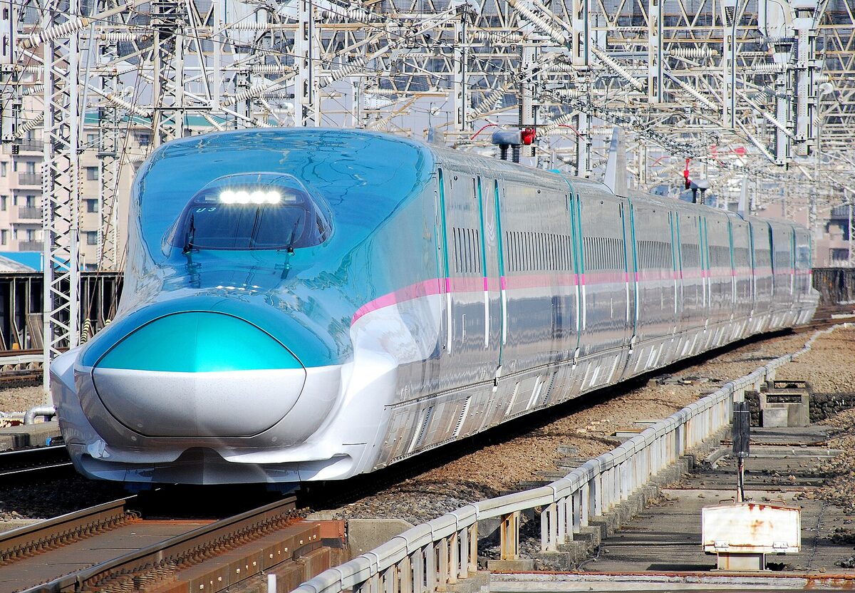 Shinkansen E5 verde turchese avanza sotto una fitta rete di cavi e strutture elettriche presso Ōmiya.
